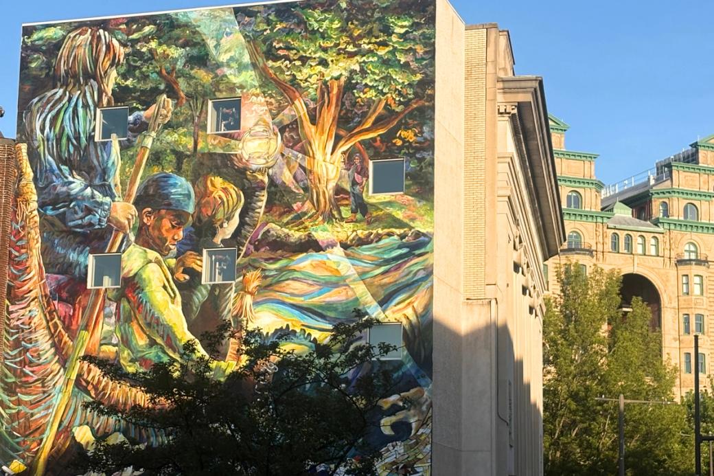 A mural on the side of a sand colored building depicts three children in a small boat. The front child holds a small glowing orb in hand that lights the way. A grove of trees surrounds the rushing water, and an older woman stands on the banks further down the waterway carrying a lamp. 