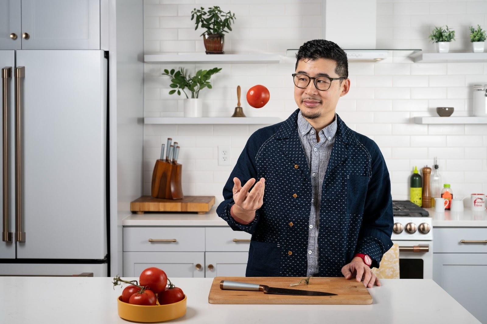 A man throwing a tomato in the air as if it were a ball. He's in the kitchen with a kitchen knife and wood block