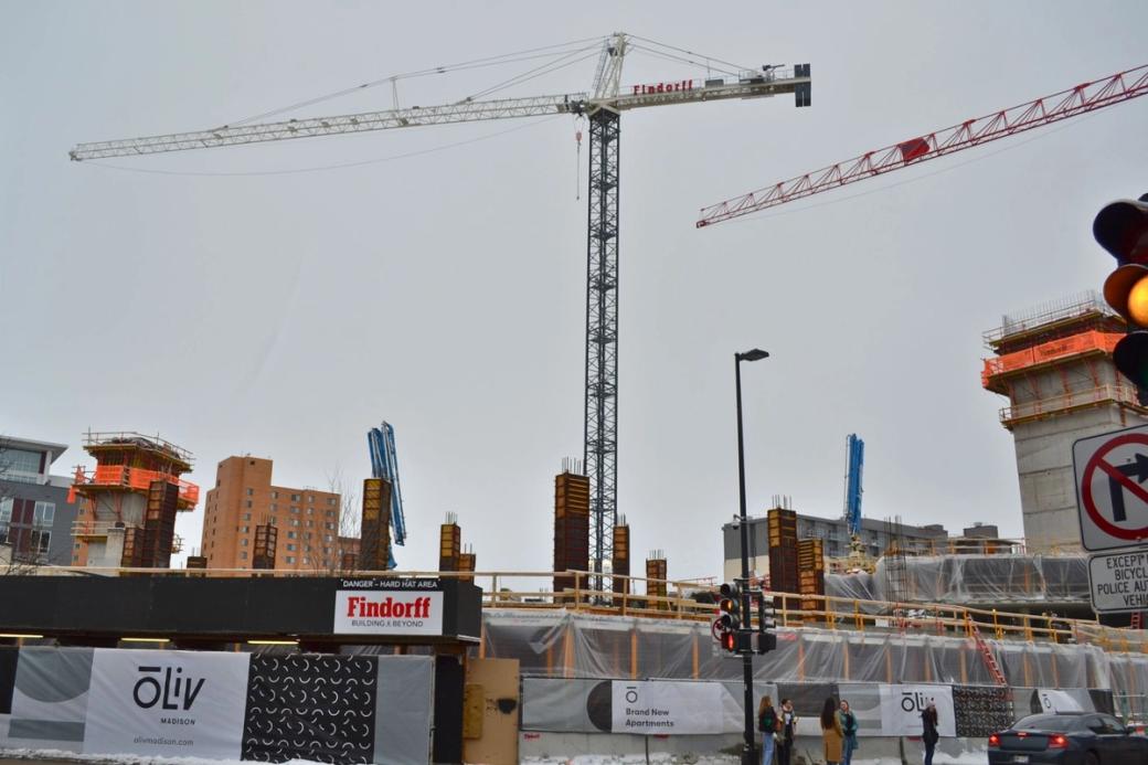 The construction site for Ōliv Madison apartments with a tall tower crane, concrete columns, and advertising banners.