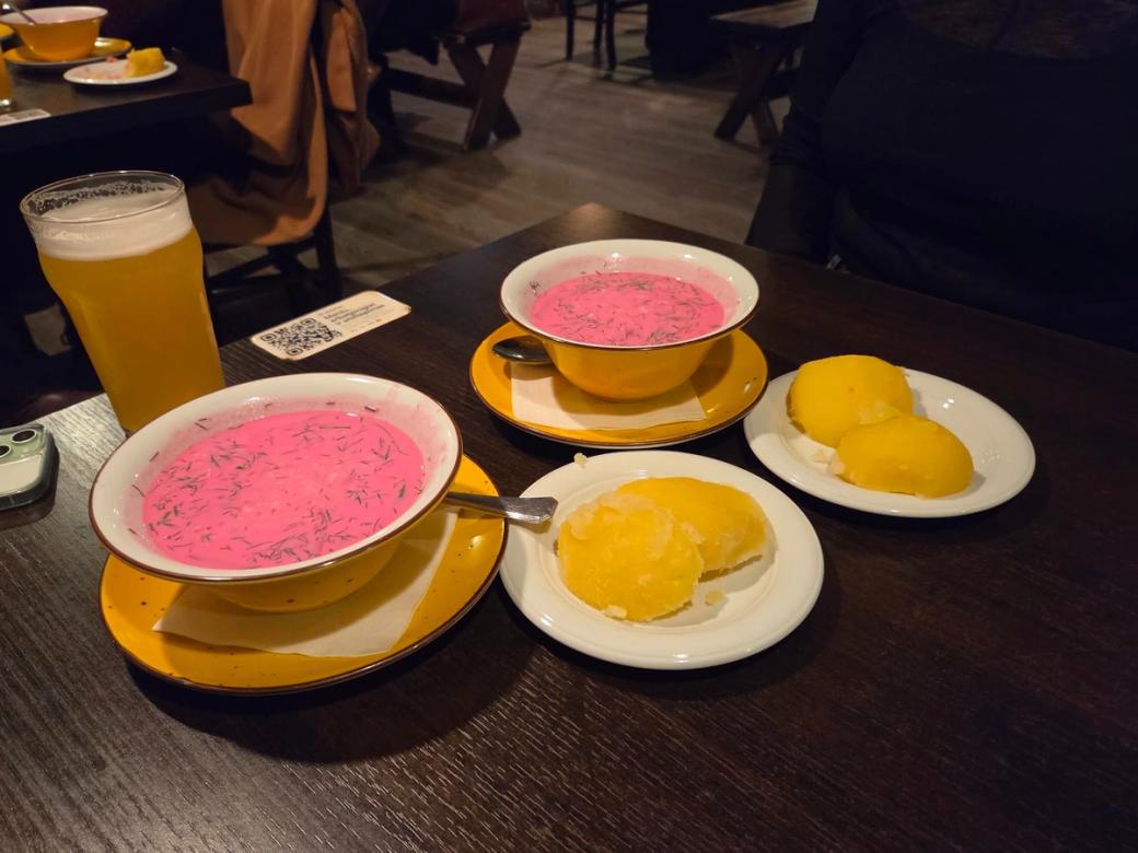 A table at a restaurant with two bowls of bright pink soup, each served on a yellow saucer, alongside plates of yellow sorbet or dessert and a glass of beer.