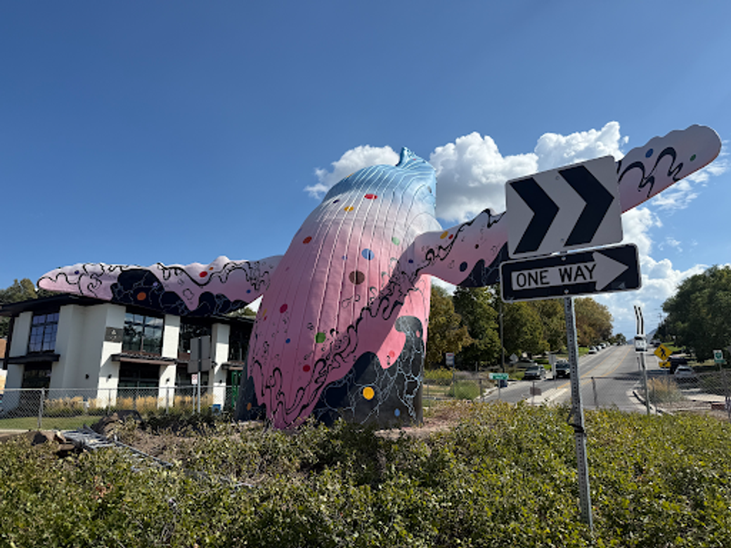 The 9th & 9th whale sculpture with its new mural, “Open Your Eyes,” arching out of a roundabout under a blue sky. The whale is painted in a pink-to-blue gradient with multicolored dots and black swirling wave patterns. 