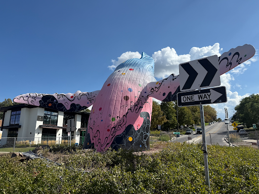 The 9th & 9th whale sculpture with its new mural, “Open Your Eyes,” arching out of a roundabout under a blue sky. The whale is painted in a pink-to-blue gradient with multicolored dots and black swirling wave patterns.