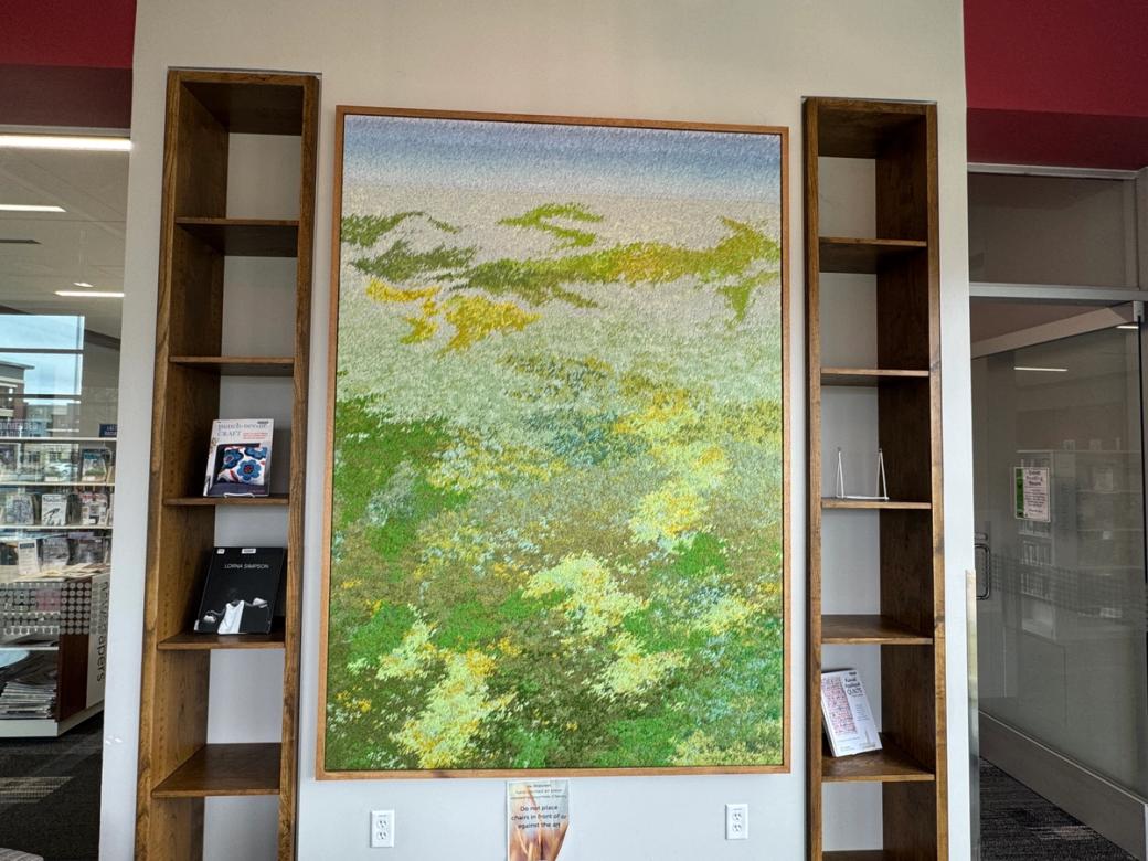 A large, rectangular artwork in the foreground with patches of all different shades of green, with a light blue band at the top. Next to and behind the artwork are some books on shelves.