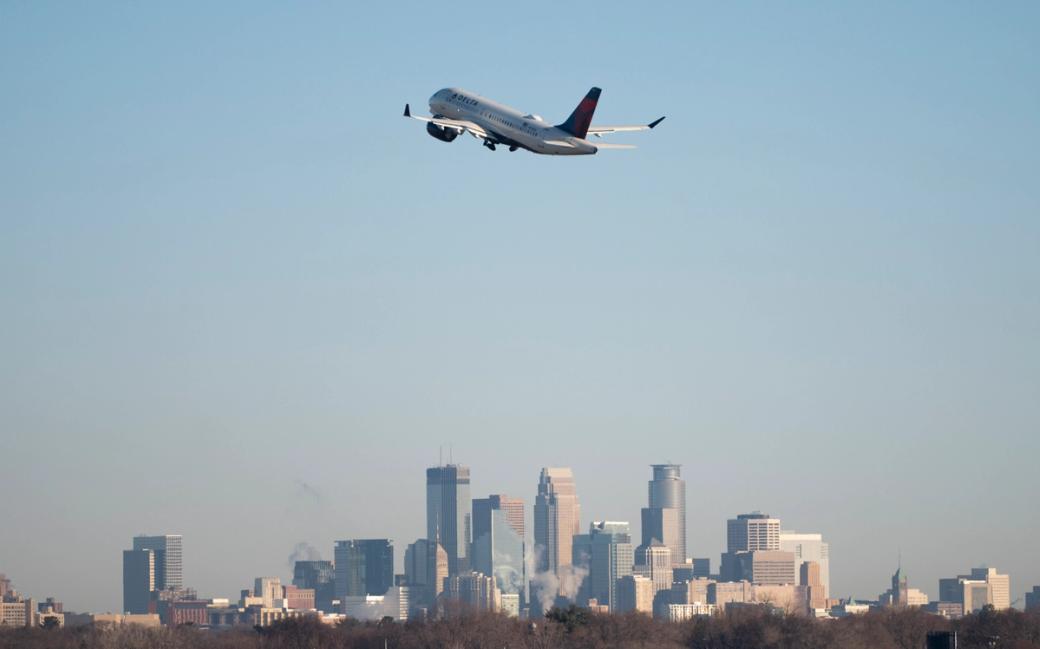 An airplane takes off into a blue sky with the Minneapolis skyline in the background.