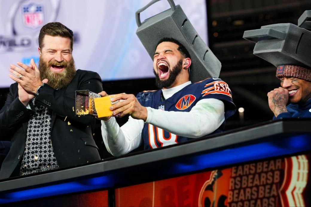 Bears quarterback Caleb Williams shreds cheese, in uniform, while wearing an oversized cheese grater style hat. Two people are next to me, one of whom is applauding, and everyone is laughing.