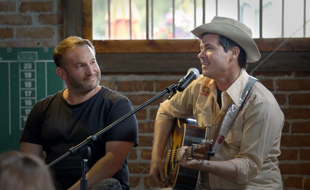 Two white men sitting on a stage. The one on the left wears a black shirt and is smiling at the one on the right, who holds a guitar and speaks into a microphone. He wears a tan button-up and a tan cowboy hat.