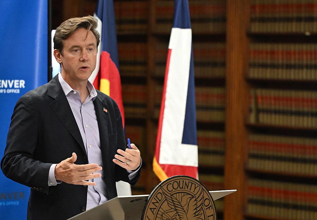 Mayor in suit stands giving a speech to constituents with flags and bookshelf in background
