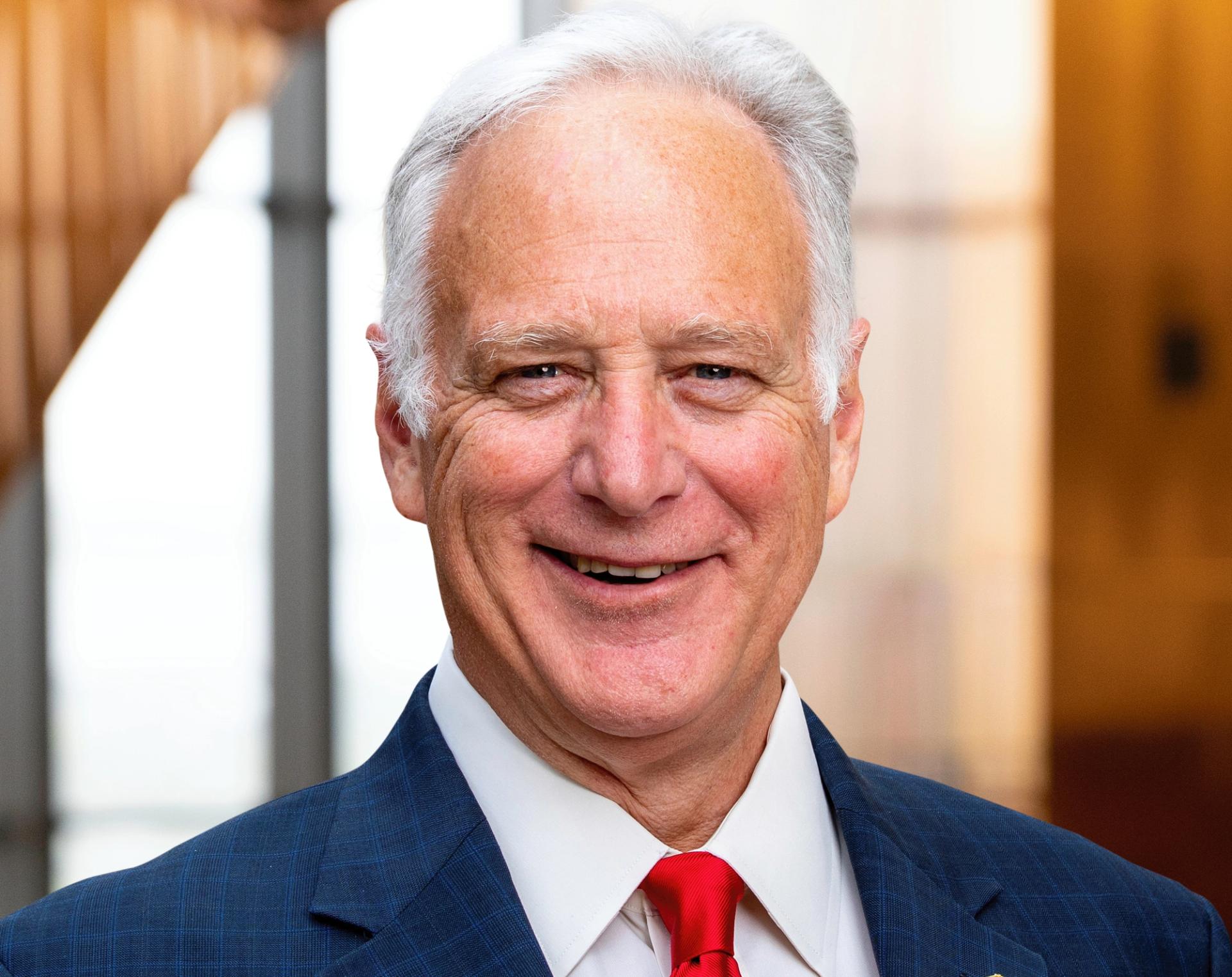 A man in a blue suit with a red tie and gray hair smiles at the camera.