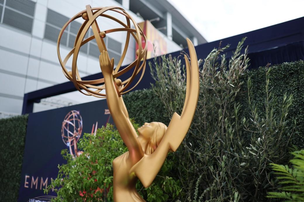A golden statue of a woman with wings holding up a globe is in the foreground, with an Emmy Awards sign and foliage in the background.