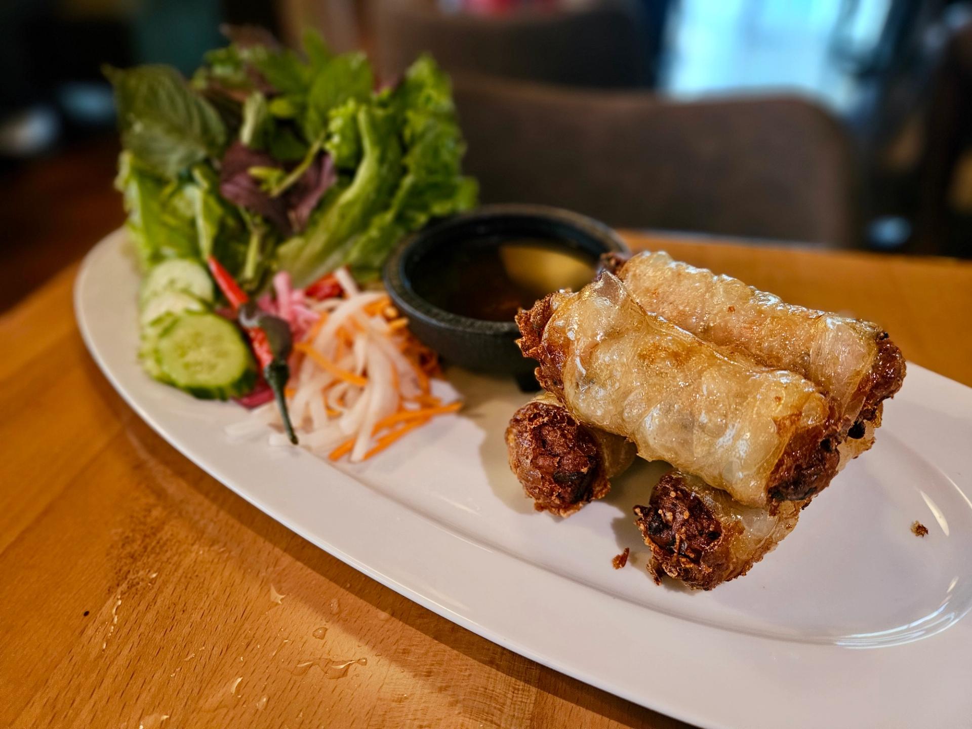 A white plate of spring rolls, garnished with some slices of orange and leaves of lettuce.