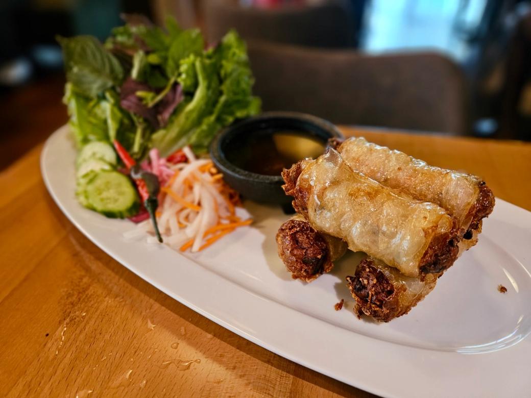 A white plate of spring rolls, garnished with some slices of orange and leaves of lettuce.