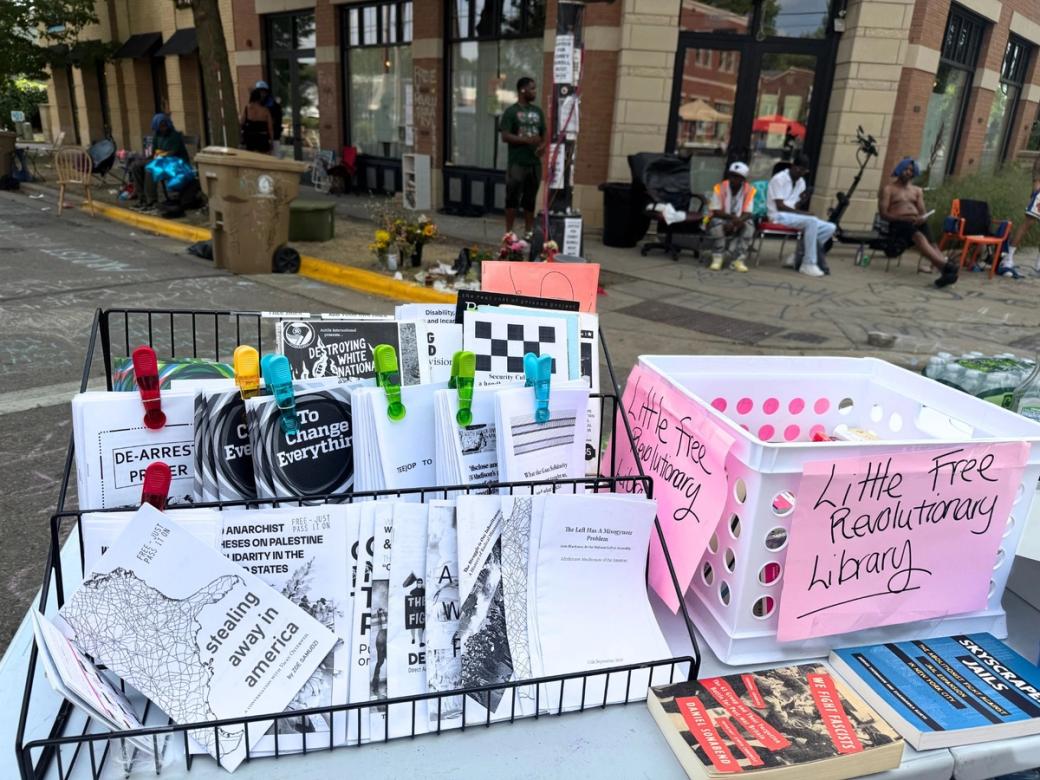 A table holds stickers and leaflets and a box that reads "Little Free Revolutionary Library."