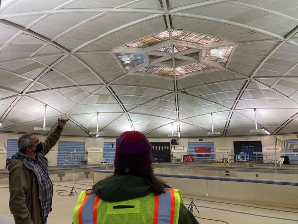A man in a mask shows off the vaulted ceilings of the Columbia Pool to a person in a construction vest and hat.