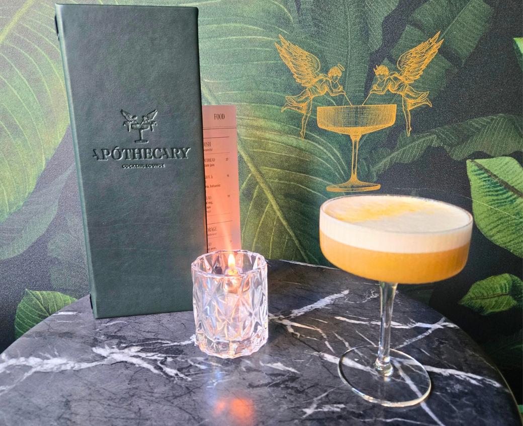 An orange cocktail with foam on top next to a candle and menu. The wallpaper behind it has banana leaves and an image of two angel babies drinking out of a glass that looks like the cocktail glass
