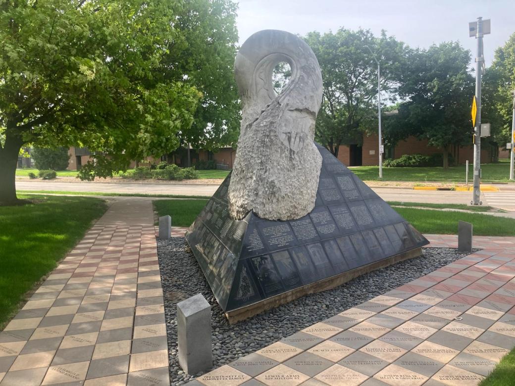 A cement snake sculpture sits atop a black pyramid on a city square.