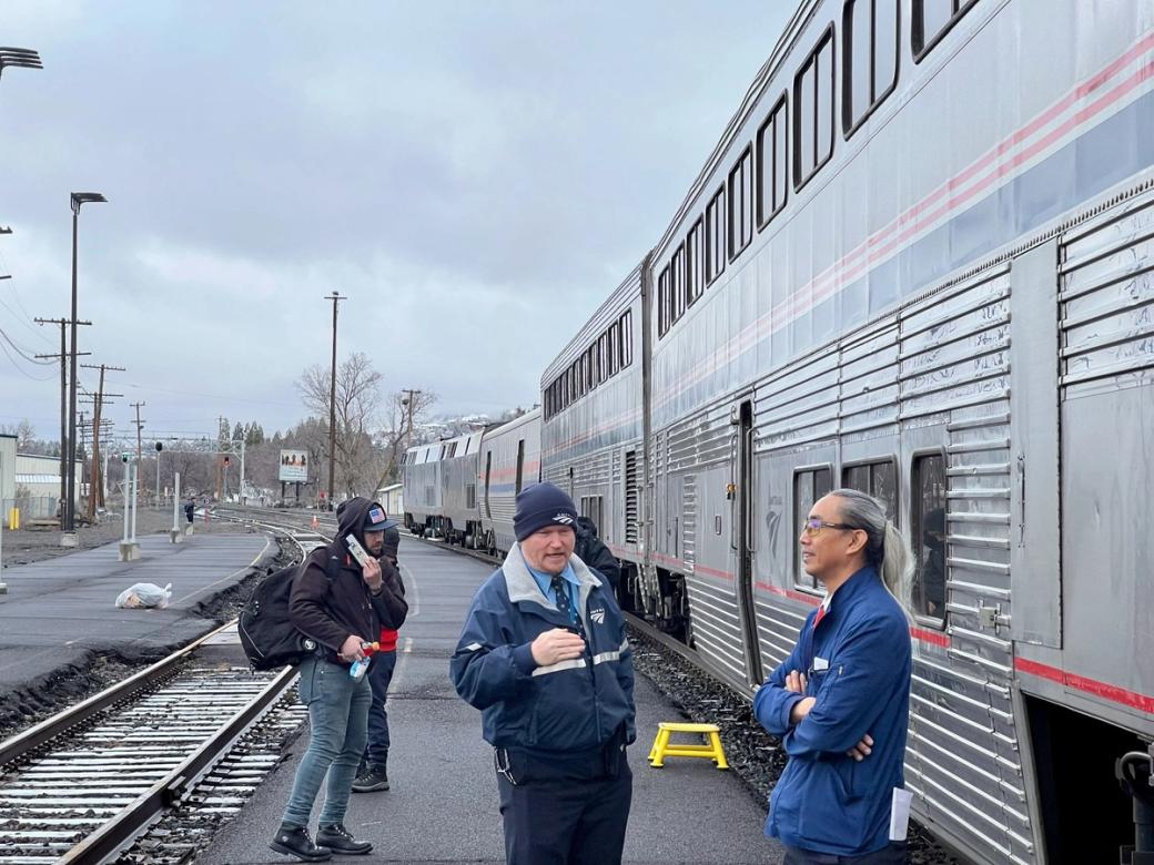 A few people standing outside of a long silver Amtrak train with red and blue stripes. 