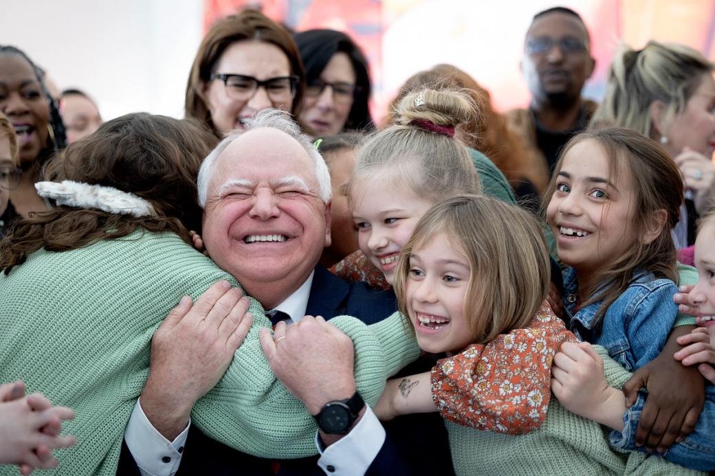 Governor Tim Walz wearing a suit, is surrounded by several hugging children. 
