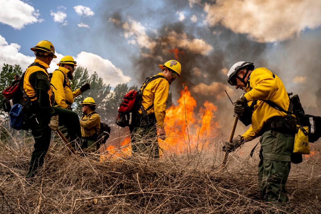 A crew of wildland firefighters in yellow gear and hard hats work alongside active flames and billowing smoke in a dry brushy landscape.