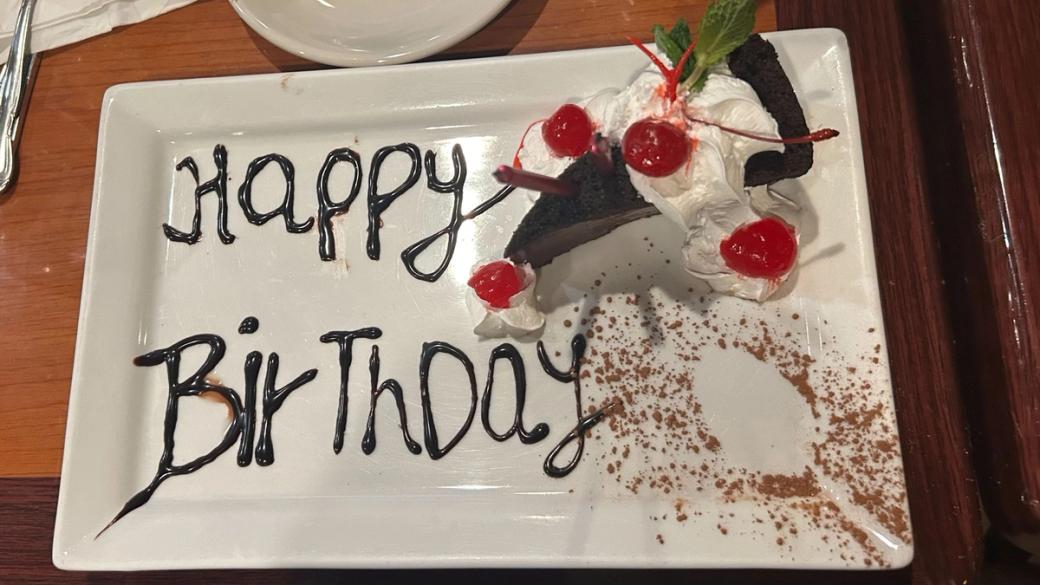 A slice of chocolate cake sitting on a white plate with "Happy Birthday" written on the plate in chocolate sauce.