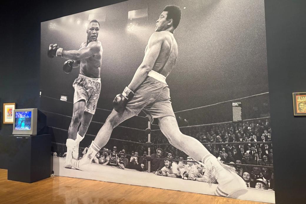 Smokin' Joe Frazier takes a swing at Muhammad Ali in a giant black and white photo on a black wall in. To the left and right other art pieces are on the walls and on a small television in the front left of the photo. 