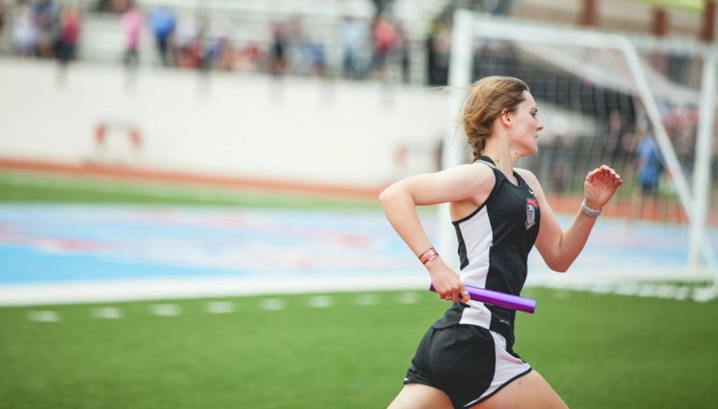 Student athlete in black and white jersey and shorts runs past the camera holding a purple baton. She has blond hair braided back and her arms are raised mid-stride