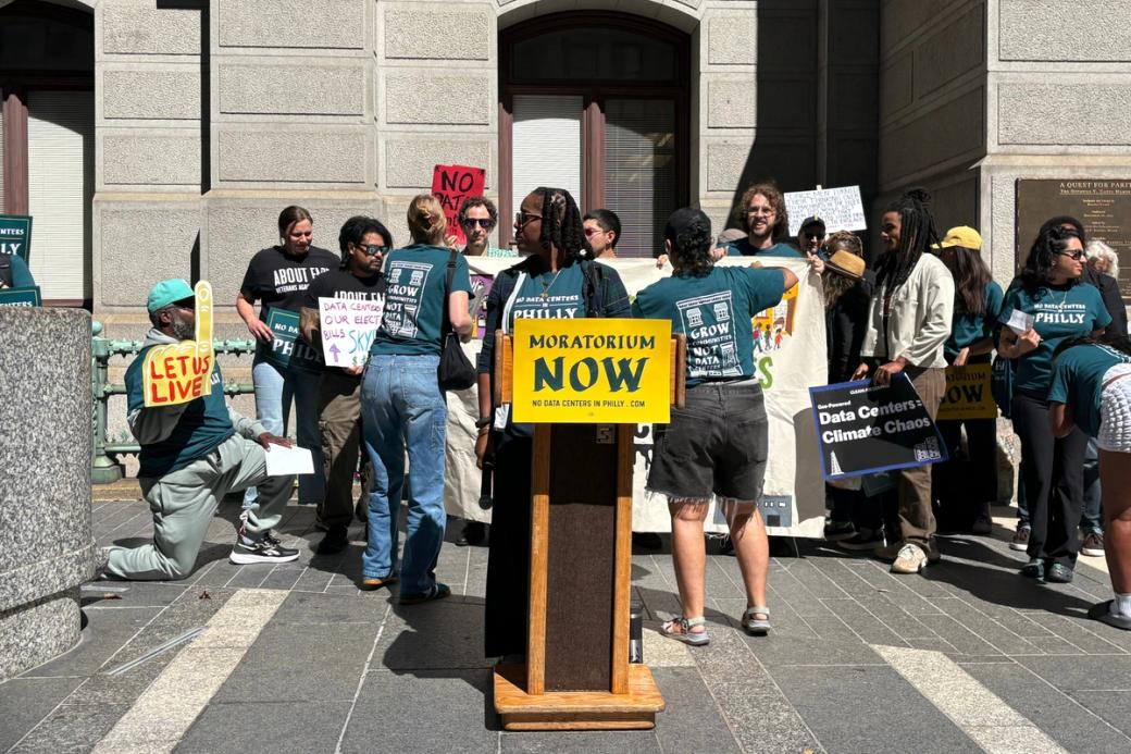 Organizers stand around a wooden podium on the southern side of City Hall. One person with short dark hair is getting ready to speak, while others gather behind in support of the “No Data Centers in Philly” campaign, holding signs like “Let Us Live,” and “Data Centers = Climate Chaos.”