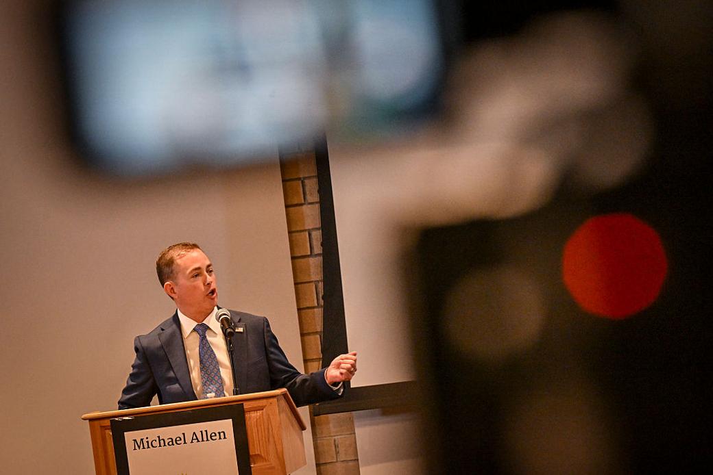 A man in a dark suit gestures with his right hand while speaking on a microphone.