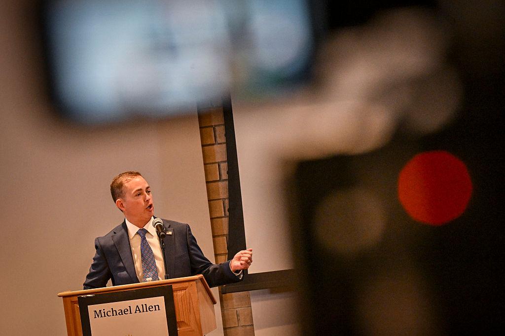 A man in a dark suit gestures with his right hand while speaking on a microphone.