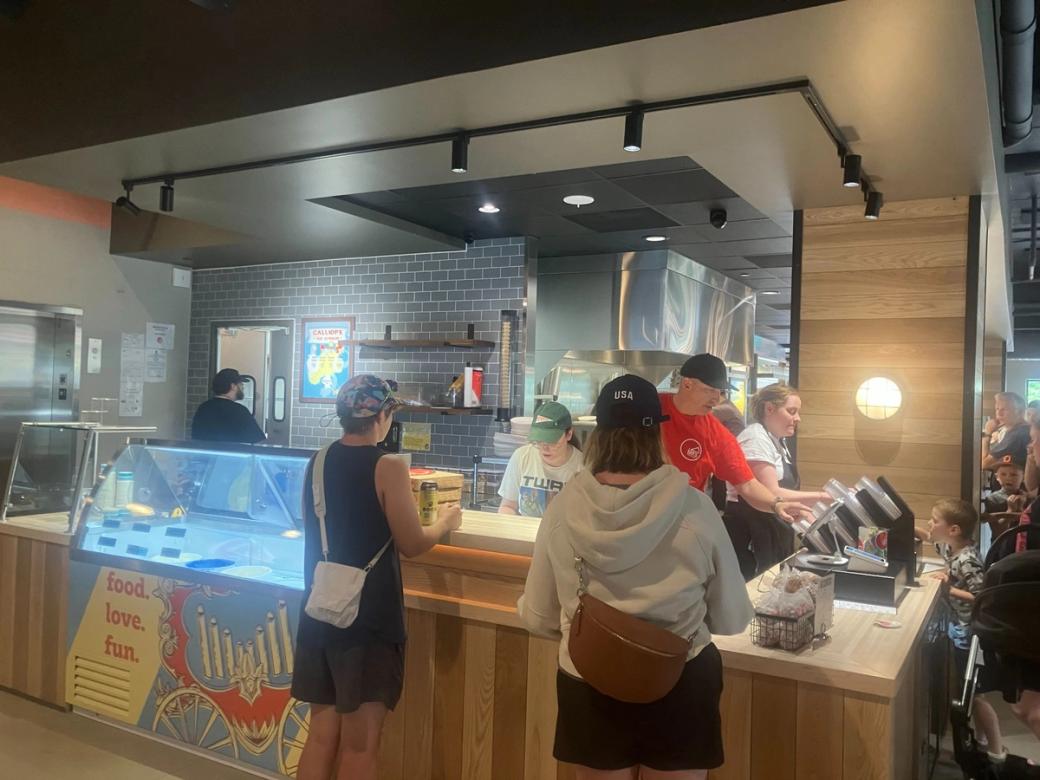 Two customers stand at the counter of Ian's Pizza. A man in a red shirt prepares food.