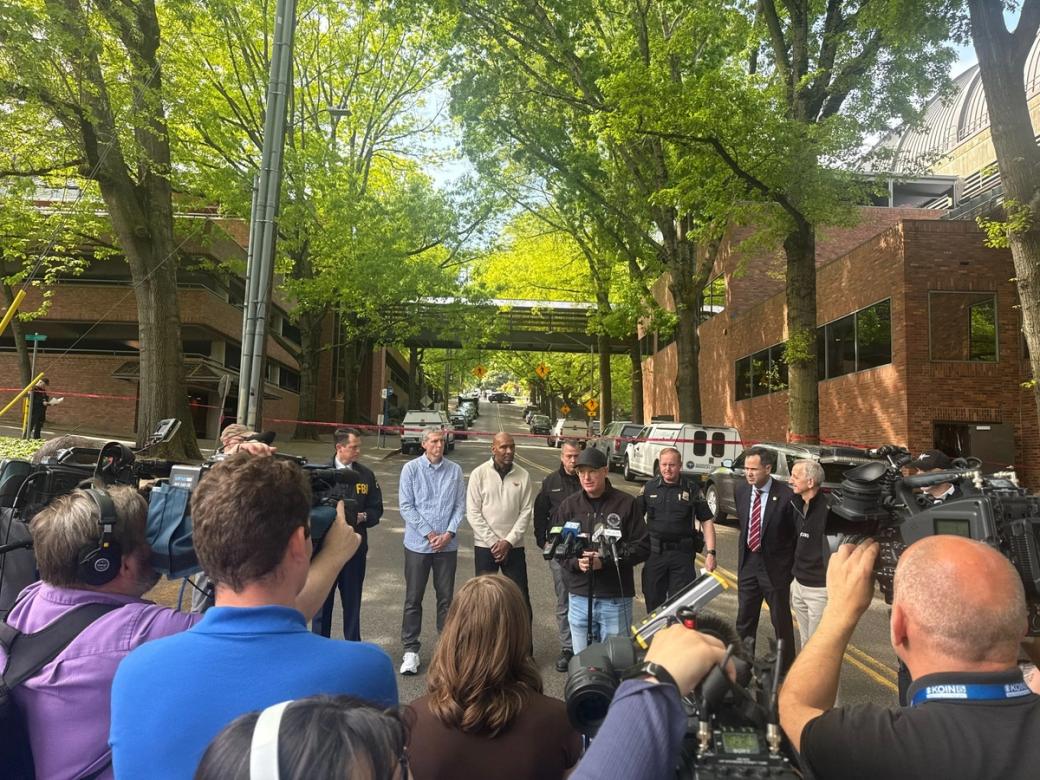 A group of reporters film a group of officials including one person in a police uniform, one person wearing an FBI jacket, the mayor of Portland and more.