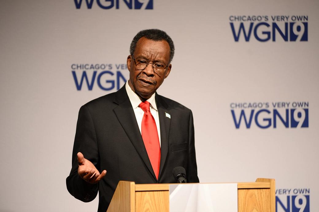 A man in a black suit and red tie speaks at a podium in front of a WGN9 step-and-repeat backdrop during a televised event.