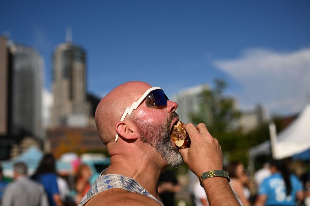 A bald person in a tanktop shoves a burger into their mouth.