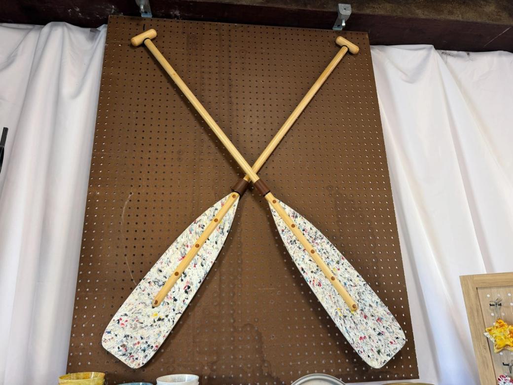 Two kayak paddles are mounted on the wall in an “X” formation. The paddle blades are white with colors speckled throughout.