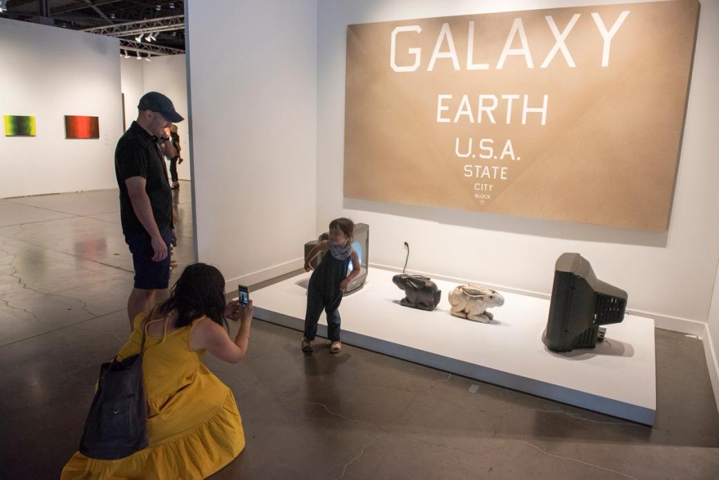 A woman in a yellow dress crouches down to take a photo of her child posing in front of an art installation with a huge brown canvas on the wall and white lettering reading “Galaxy, Earth, U.S.A., State, City” in descending order and a few electronics and sculptures scattered on the floor below.