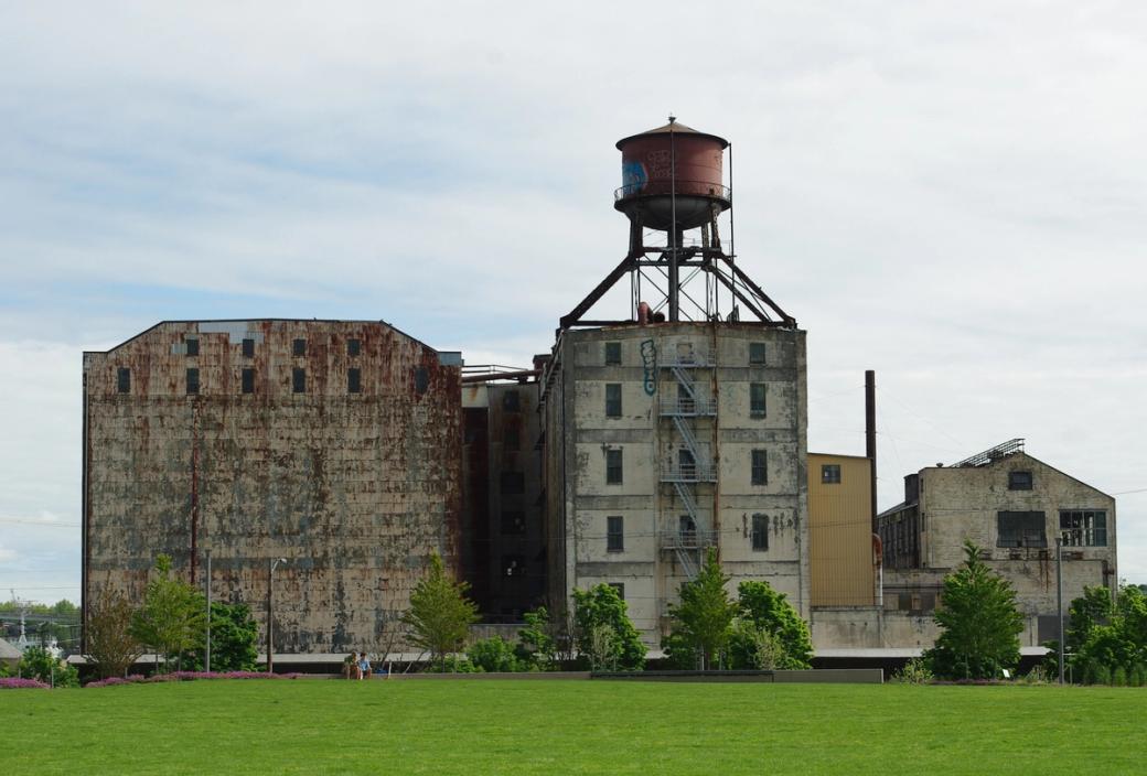 Large industrial building with water tower on top, looks to be in disrepair. Viewed from a distance on a cloudy day with grassy area in foreground.