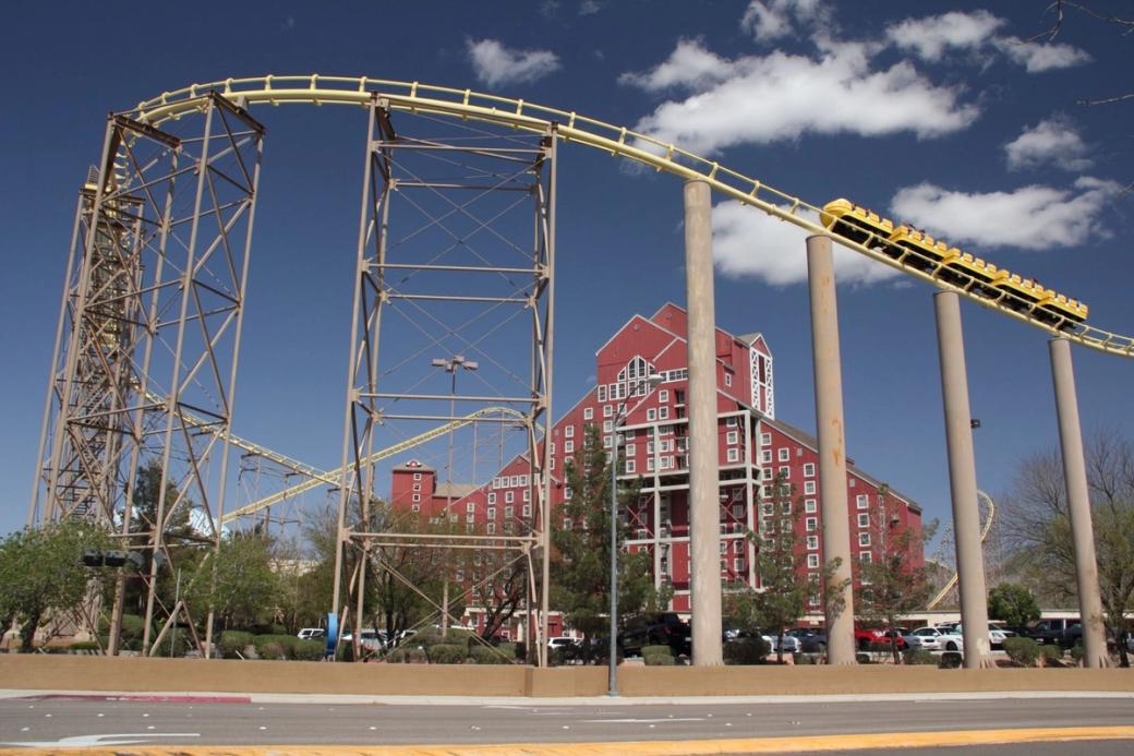 Yellow roller coaster train descending a track past a large red multi-story building under a blue sky with white clouds.