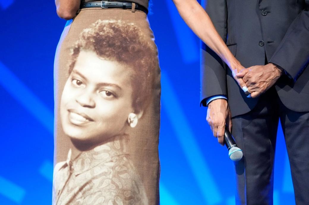 The bottom half of a woman and a man on a stage with a mic. The woman has an old photo of a woman printed on her dress