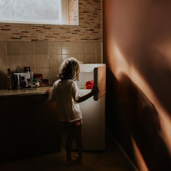 In a darkened kitchen, a small child aged perhaps 7 or 8, holds open a freezer door. The inside is mostly bare, aside from a plastic-wrapped loaf of bread, and one other small package.