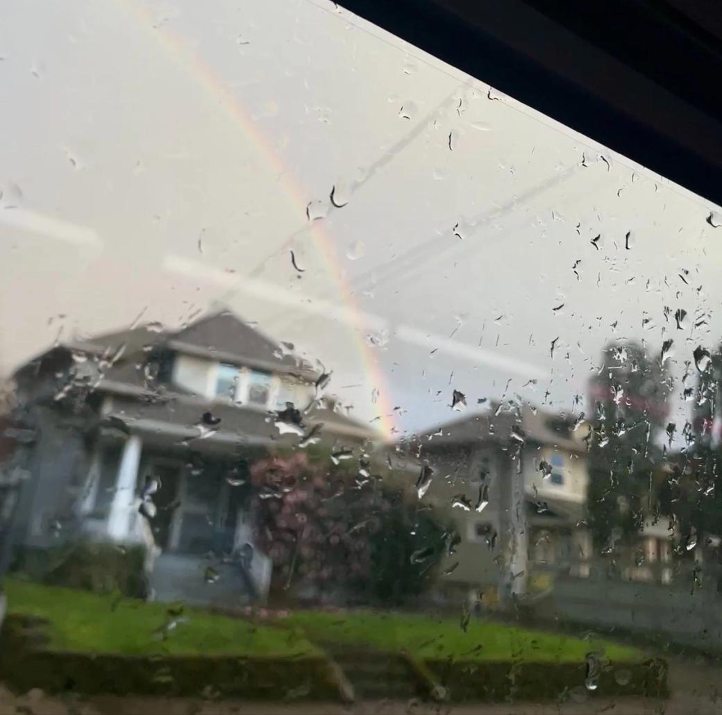 A view from a rain-covered window with houses, grass, and trees in the distance and a rainbow in the cloudy sky above.