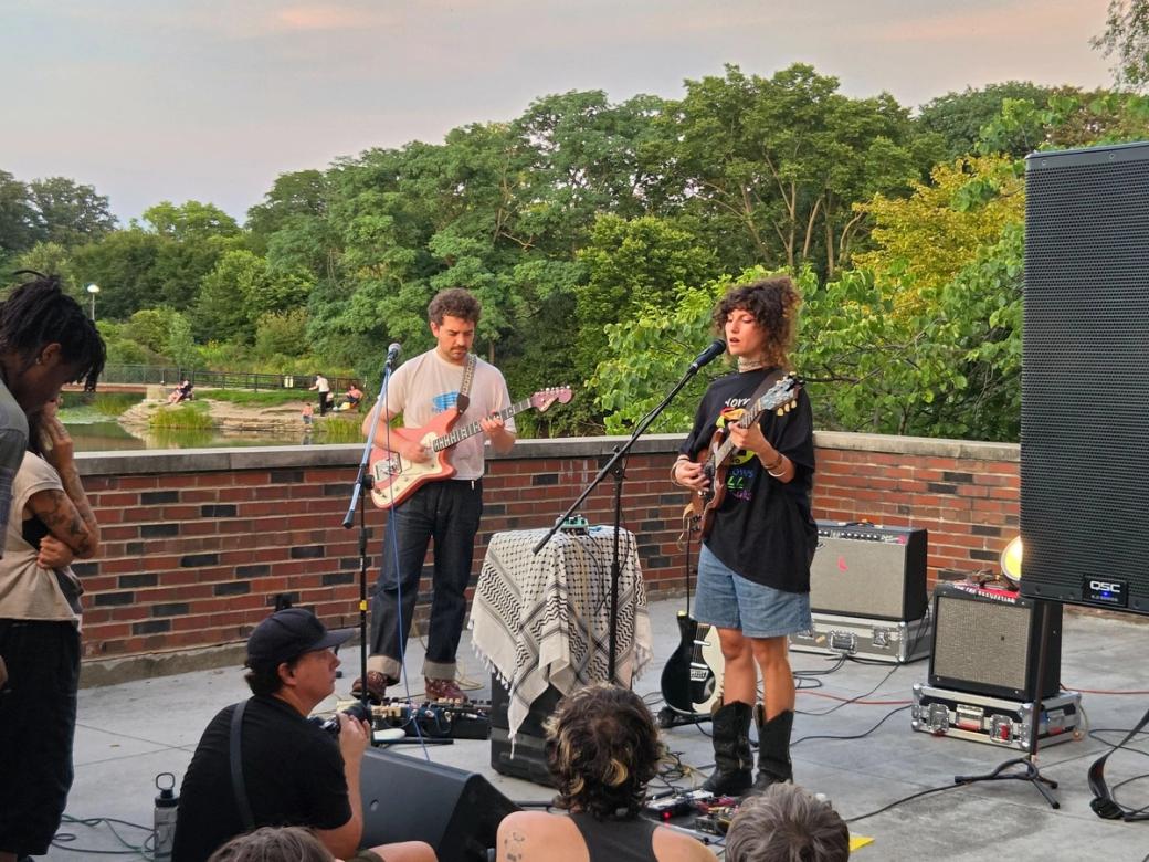 Two people, one man, one woman, playing guitars and singing in a mic in front of a small crowd