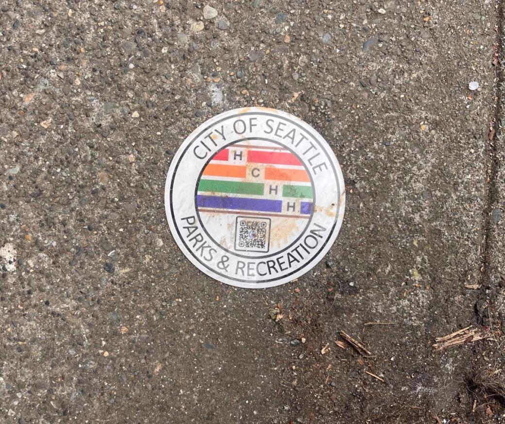 A round white disk with four colorful stripes in red, orange, green and blue, the letters H, C, H, H and a QR code and the name of Seattle Parks and Recreation in the outer circle is placed on the sidewalk. It looks a bit dirty and like it’s been stepped on.