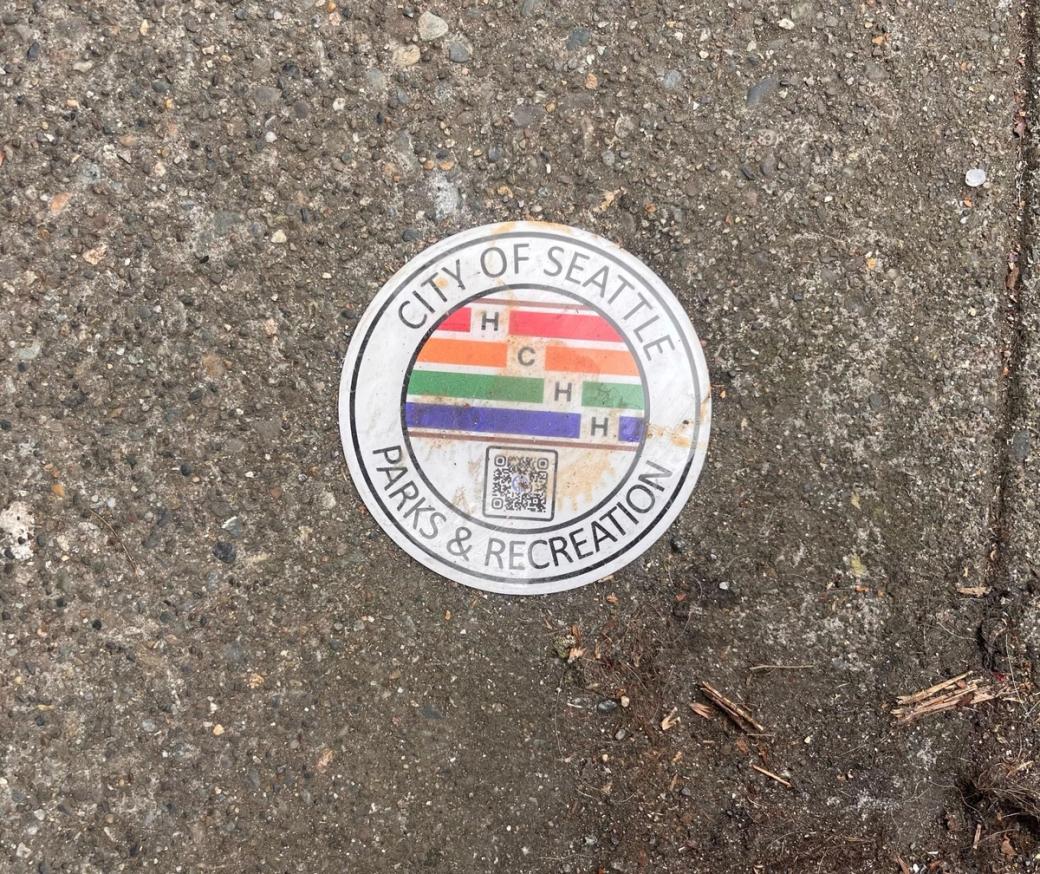 A round white disk with four colorful stripes in red, orange, green and blue, the letters H, C, H, H and a QR code and the name of Seattle Parks and Recreation in the outer circle is placed on the sidewalk. It looks a bit dirty and like it’s been stepped on.