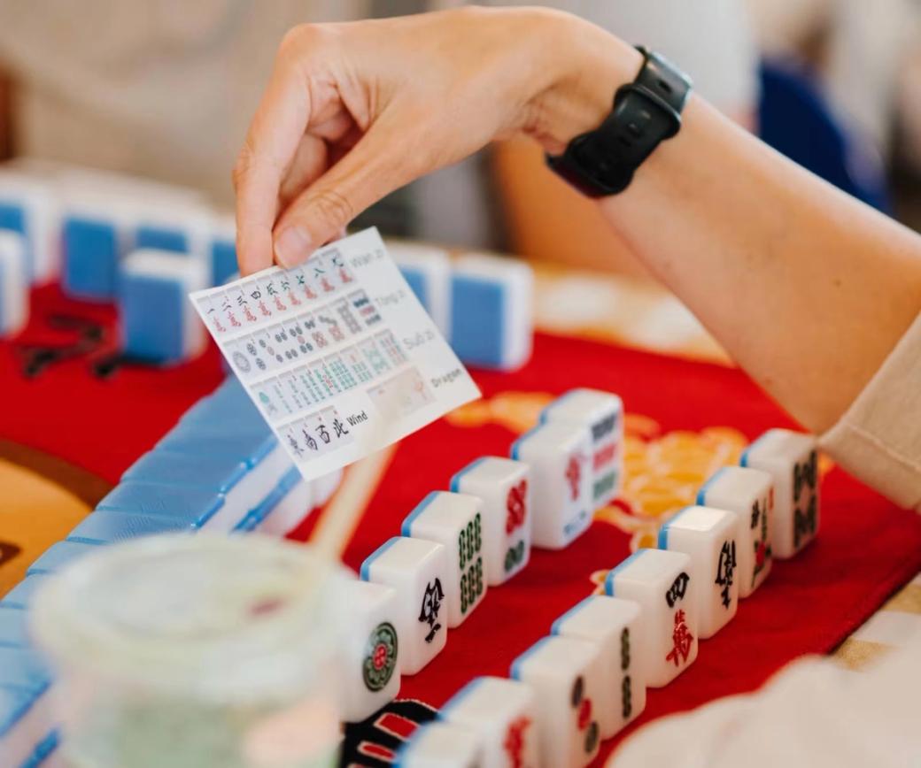 Mahjong tiles sit on a table, and a hand holds a card with images of tiles over them.