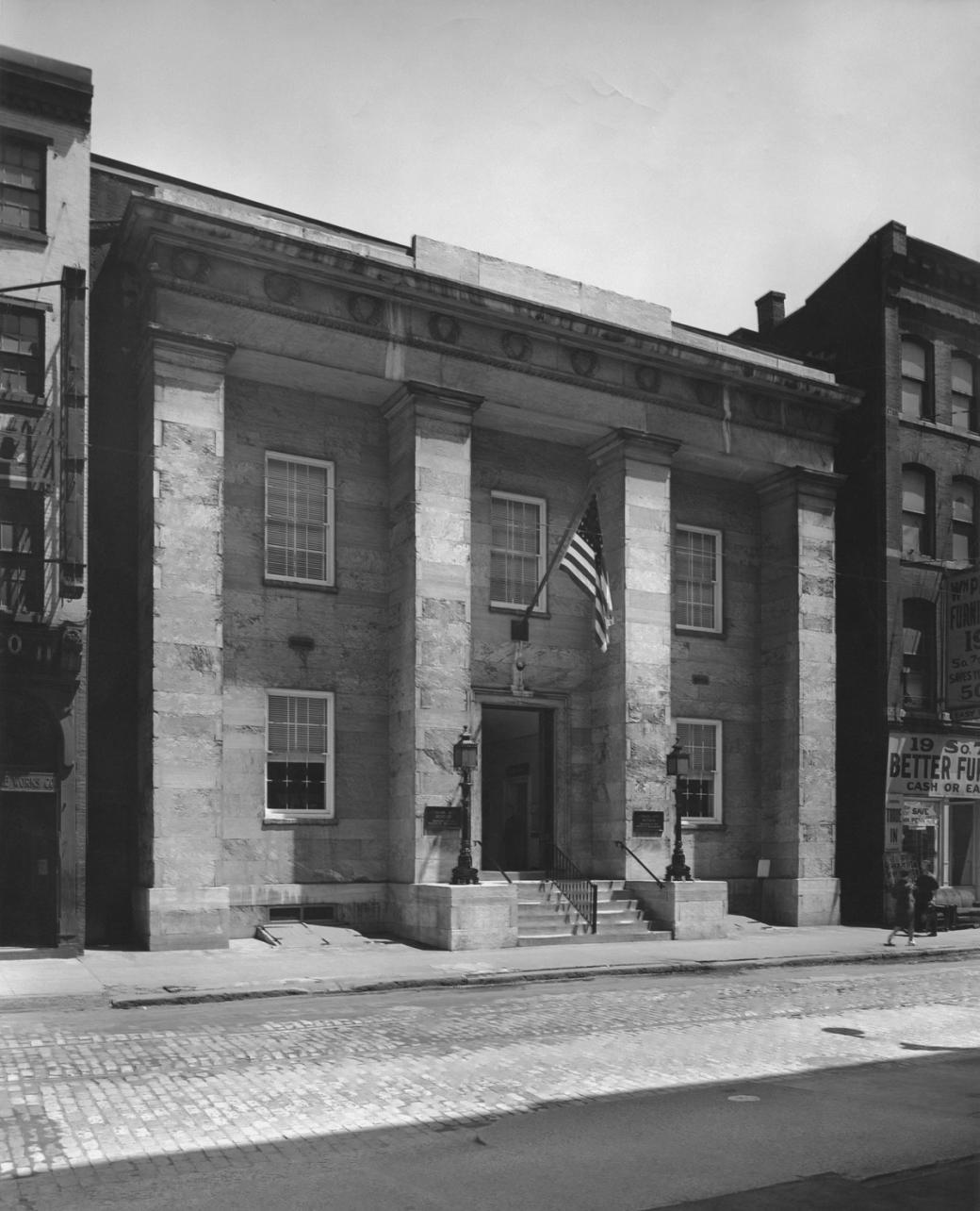 A black-and-white photo of the two-story building, constructed in the Classical Greek Revival design with pillars. An American flag hangs above the door.
