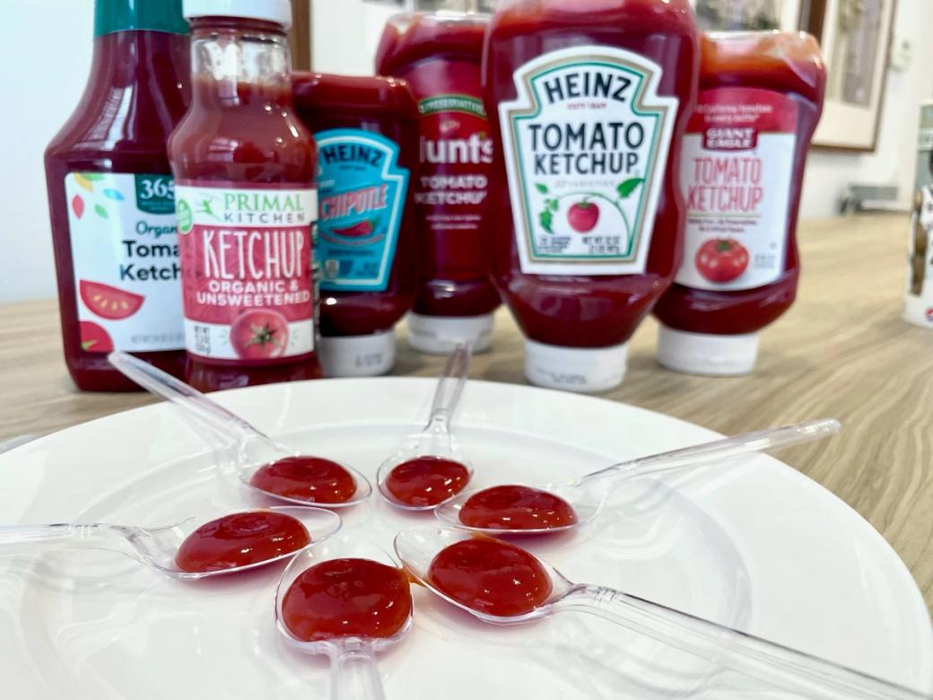 Six plastic spoons in a circle, each holding a dollop of ketchup from a different bottle.