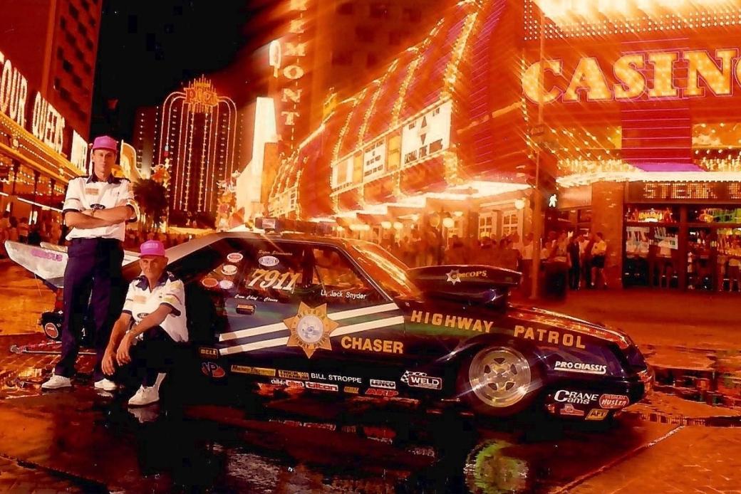 Two men pose on the Strip in front of a black car that says "Highway Patrol" and "Chaser".