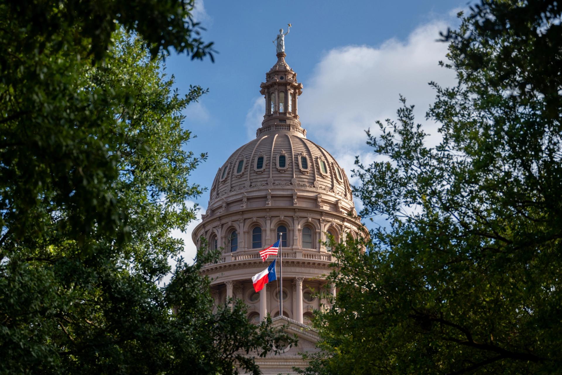 The outside of the Texas Capitol surrounded by green trees. A Texas flag and a U.S. flag are hanging outside.