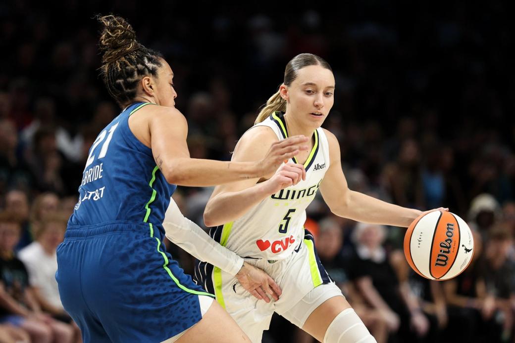 Two women wearing different team jerseys playing basketball, with large out of focus crowd in background