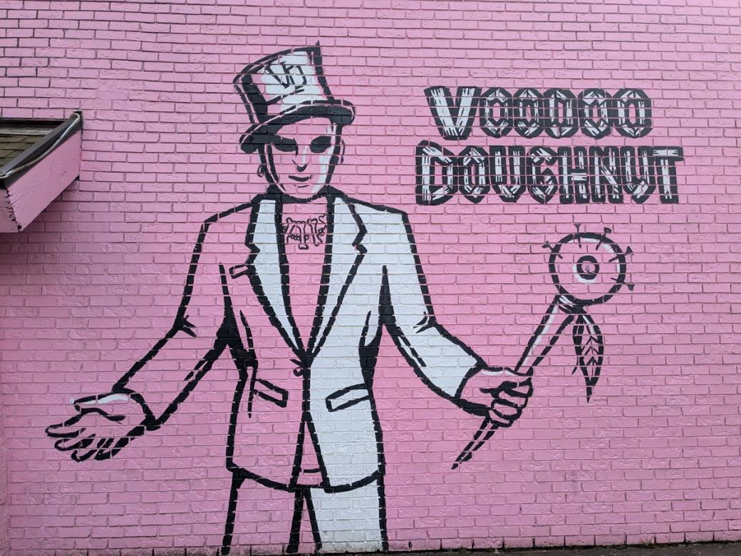 pink brick wall with mural of man holding a doughnut on a stick with words "Voodoo Doughnut"