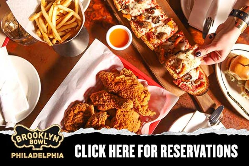 A table with plates of crispy fried chicken, fries, pizza slices, and a burger. A hand reaches for a slice. Text reads "Brooklyn Bowl Philadelphia."